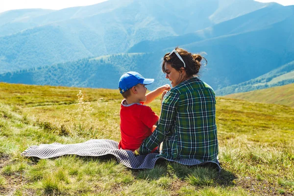 Madre joven con un niño sentado en una manta en la cima de la montaña. hermosa vista del paisaje en el fondo —  Fotos de Stock