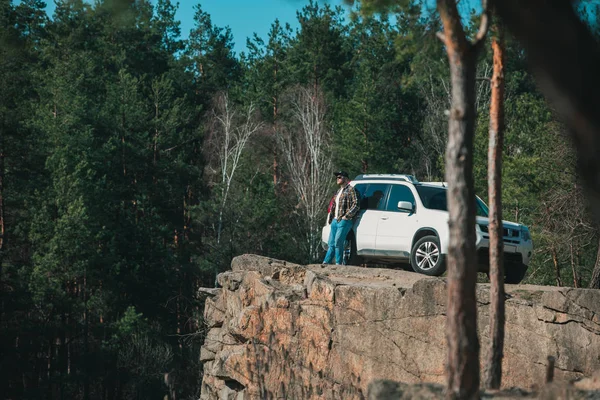 Людина стоїть на скелі скелі біля білої позашляховик автомобіль з видом на озеро. Перегляд дерев — стокове фото