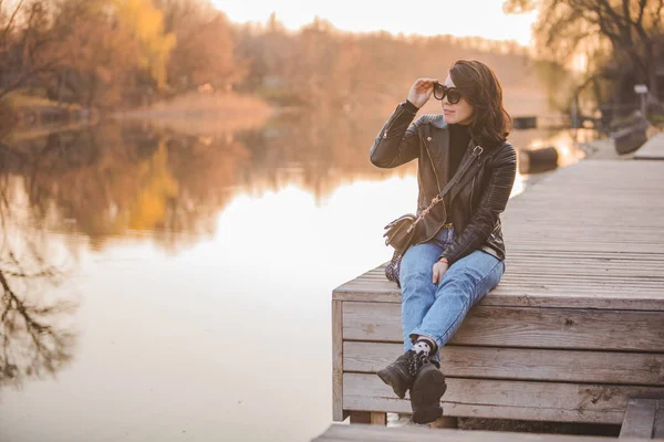 Giovane bella donna di moda seduta sul molo al tramonto con bella vista sul fiume — Foto Stock
