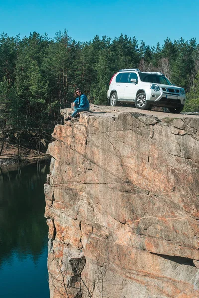 Mujer sentada en el borde rocoso con vista al lago. coche suv blanco cerca — Foto de Stock