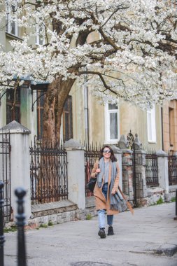 young stylish woman walking by city street in coat blooming tree on background