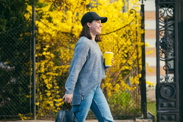 Young stylish woman in cap drinking coffee outdoors from yellow paper cup — Stock Photo, Image