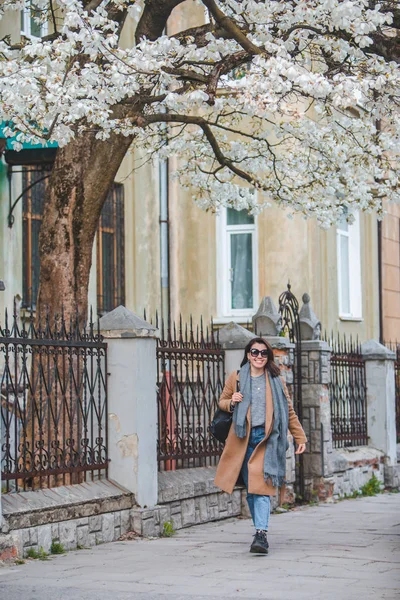 Young stylish woman walking by city street in coat blooming tree on background — Stockfoto