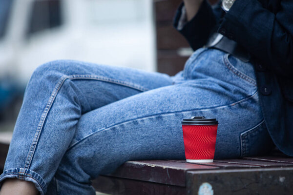 red paper coffee cup at bench near woman legs in blue jeans
