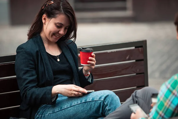 Mulher bebendo café no banco da cidade olhando para o telefone — Fotografia de Stock