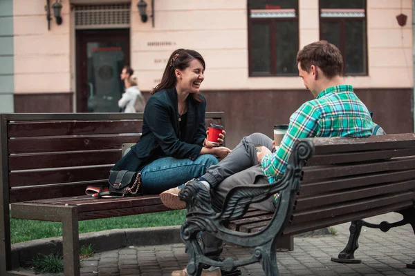 Riendo hablando hombre con mujer sentada en el banco bebiendo café navegando en internet. estilo de vida urbano —  Fotos de Stock
