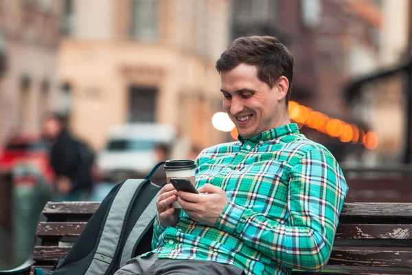 Retrato de jovem sorrindo bonito homem sentado no banco bebendo café navegando internet via telefone — Fotografia de Stock