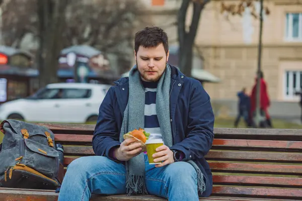 Jovem homem sorrindo comer hambúrguer e beber café sentado no banco da cidade — Fotografia de Stock