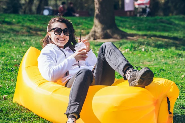 Woman drinking smoothie in city park laying on yellow inflatable mattress — 스톡 사진