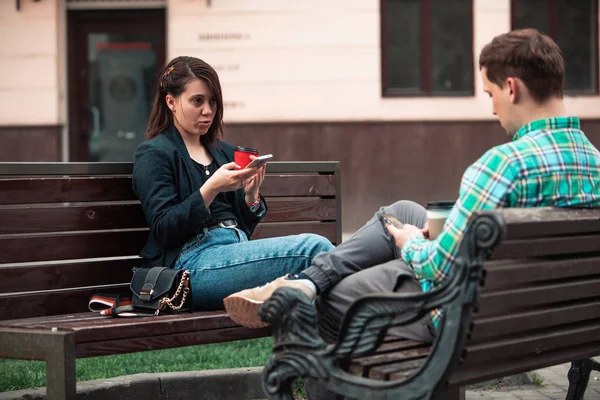 Communication problem man with woman sitting on bench at city street drinking coffee in paper cup looking ito phones — Stock Photo, Image