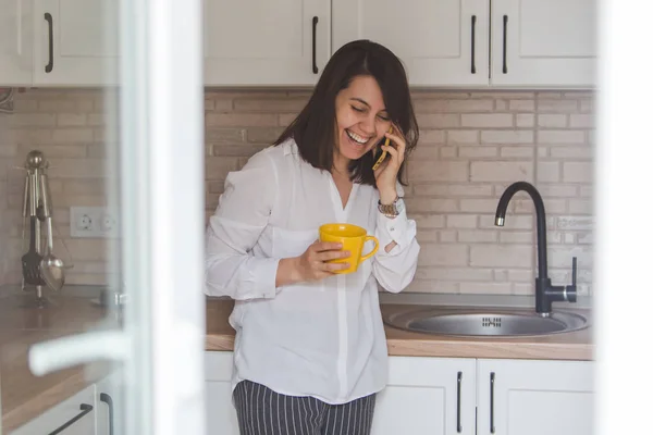 smiling happy woman talking no phone drinking tea from yellow mug at the kitchen