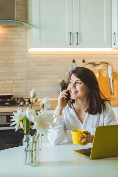 Young pretty caucasian woman talking on the phone sitting in front of laptop in the kitchen — Stock Photo, Image