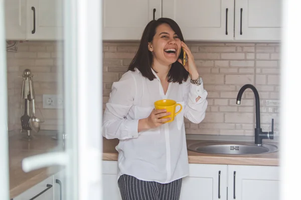 Sorrindo mulher feliz falando sem telefone beber chá de caneca amarela na cozinha — Fotografia de Stock