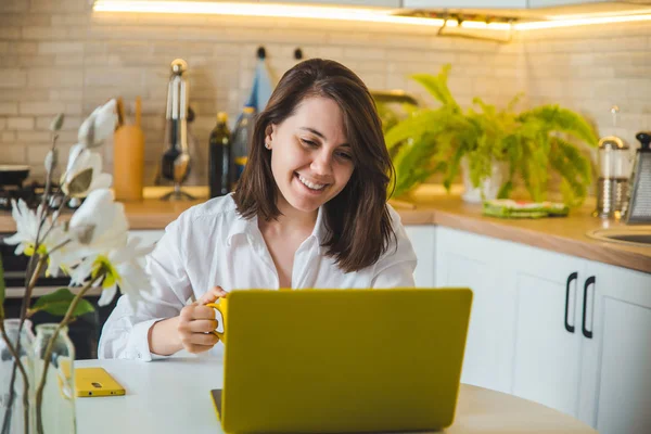 Mulher bebendo chá de caneca amarela trabalhando no laptop. cozinha no fundo — Fotografia de Stock