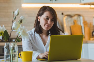 genç güzel beyaz kadın mutfakta dizüstü önünde oturan telefonda konuşurken