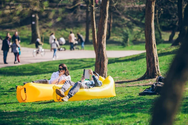 Couple laying on yellow inflatable mattress in city park. reading book. drinking smoothies — Stock Photo, Image