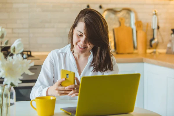 Jovem muito caucasiano mulher falando no telefone sentado na frente de laptop na cozinha — Fotografia de Stock