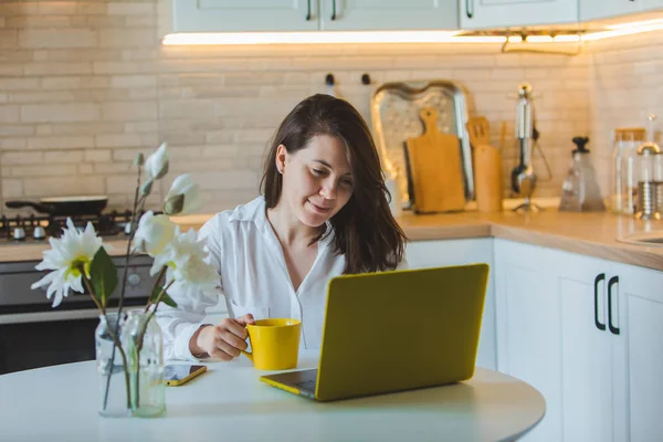 Young pretty caucasian woman talking on the phone sitting in front of laptop in the kitchen — Stock Photo, Image