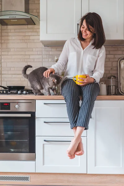 Mujer sentado en cocina mesa con gato beber té de amarillo taza — Foto de Stock