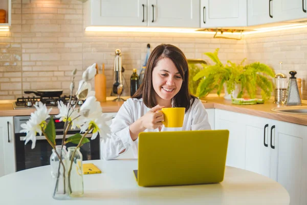 Mulher bebendo chá de caneca amarela trabalhando no laptop. cozinha no fundo — Fotografia de Stock