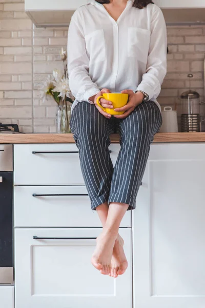 Mulher sentada na mesa da cozinha com telefone e caneca amarela — Fotografia de Stock