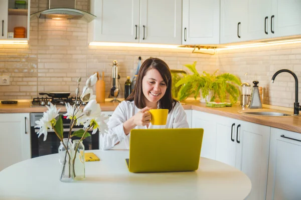 Mulher bebendo chá de caneca amarela trabalhando no laptop. cozinha no fundo — Fotografia de Stock