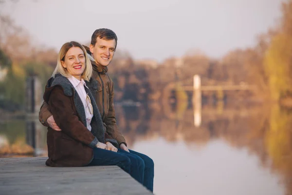 Linda pareja sentada a orillas del río al atardecer en la cálida noche de otoño —  Fotos de Stock