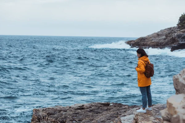 Mulher de pé no penhasco olhando no mar tempestuoso em capa de chuva amarela — Fotografia de Stock