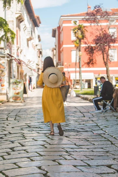 Žena ve žlutých šatech s kouskem slaměných klobouků, procházky po starém turistickém městě v Chorvatsku — Stock fotografie