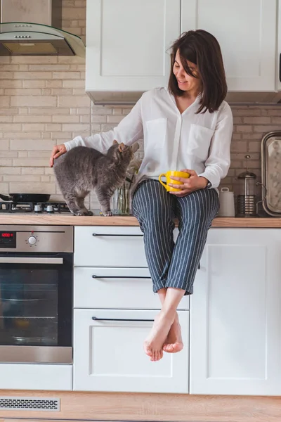 Mujer sentado en cocina mesa con gato beber té de amarillo taza — Foto de Stock