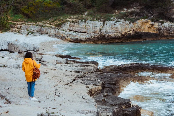 Mulher andando à beira-mar e fotografar paisagem em capa de chuva amarela . — Fotografia de Stock