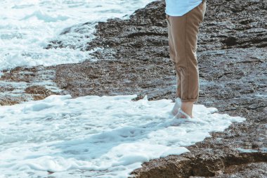 Deniz kıyısında yürüyen pantolonlu ıslak adam bacakları. Kayalık plajda suyun tadını çıkarıyor.