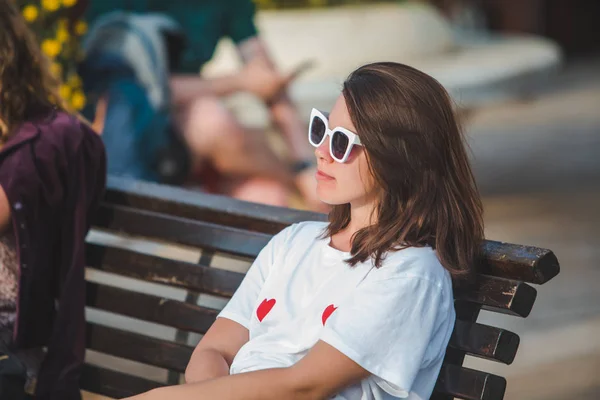 Жінка в білій футболці і білих сонцезахисних окулярах, що сидять на міській лавці — стокове фото