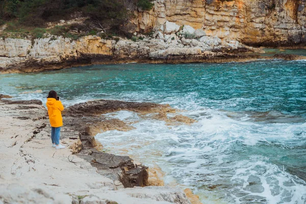 Sarı yağmurluk lu kadın kayalık plajda dikilen görmek bakıyor — Stok fotoğraf