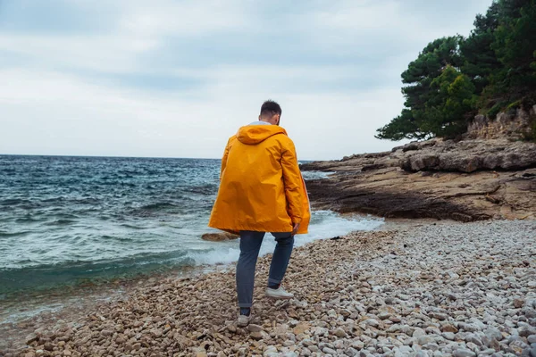 Homem andando pela praia rochosa em capa de chuva amarela. tempo ventoso nublado — Fotografia de Stock