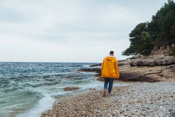 黄色いレインコートを着た岩のビーチを歩いている男。曇り風の強い天気 — ストック写真
