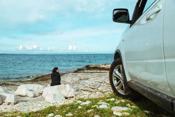 Žena sedící na skále poblíž vozu SUV u moře užívající výhled — Stock fotografie
