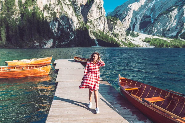 Женщина, идущая по пирсу горного озера с деревянными лодками в красном платье — стоковое фото