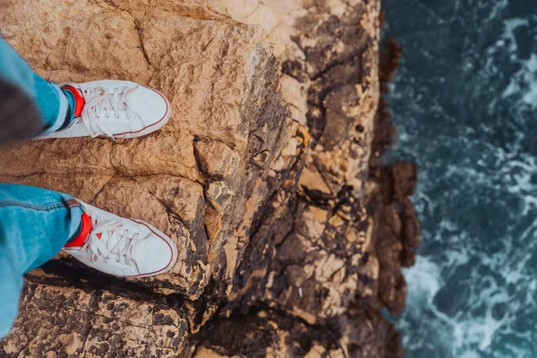 Jambes homme en jeans et baskets blanches au bord de la falaise vue sur la mer bleue orageuse — Photo