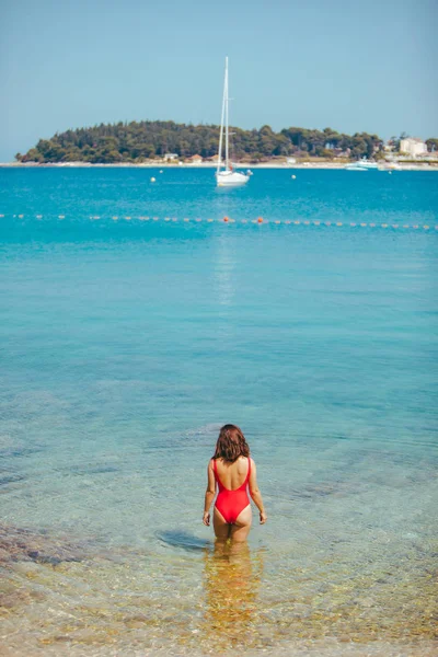 Sexy mujer en traje de baño rojo de pie en agua de mar. yate de costa en el fondo —  Fotos de Stock