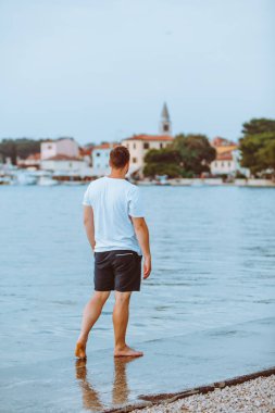 Hırvatistan 'da arka planda yaz akşamı Fazana kasabasında deniz Plajı ile yürüyen adam