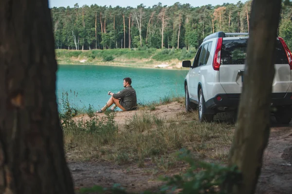Homem sentado perto de carro branco SUV na borda olhando para o lago com água azul — Fotografia de Stock