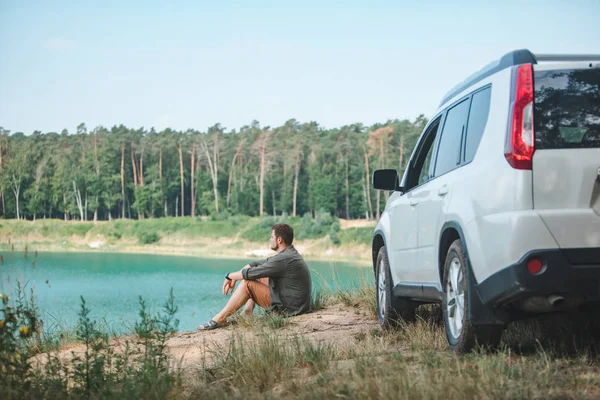 Homem sentado perto de carro branco SUV na borda olhando para o lago com água azul — Fotografia de Stock