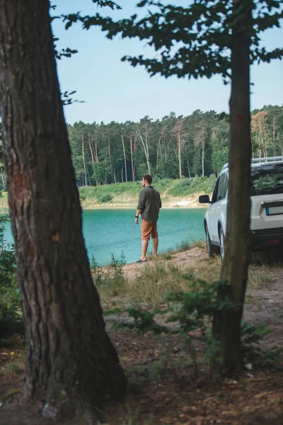 Homem perto de carro branco SUV na borda olhando para o lago com água azul — Fotografia de Stock