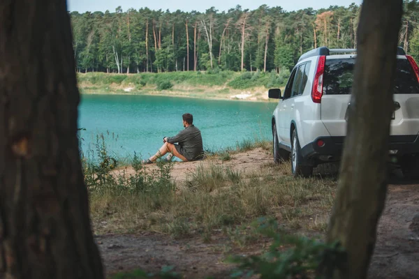 Hombre sentado cerca de coche suv blanco en el borde mirando al lago con agua azul — Foto de Stock