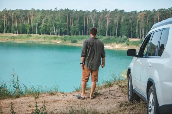 Człowiek w pobliżu białego samochodu SUV na brzegu patrząc na jezioro z błękitną wodą — Zdjęcie stockowe