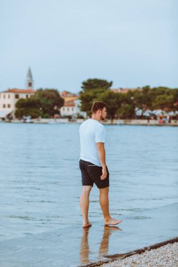 Hırvatistan 'da arka planda yaz akşamı Fazana kasabasında deniz Plajı ile yürüyen adam