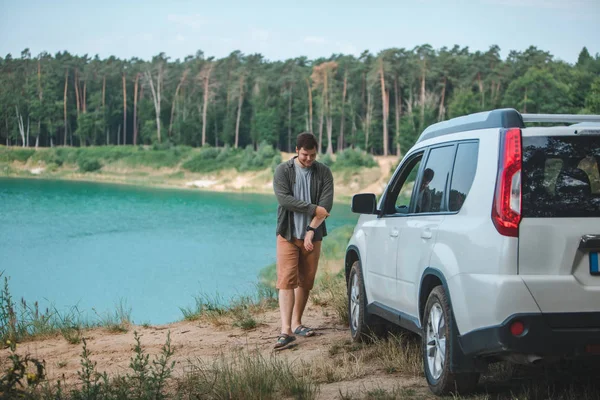 Hombre cerca de coche suv blanco en el borde mirando al lago con agua azul — Foto de Stock