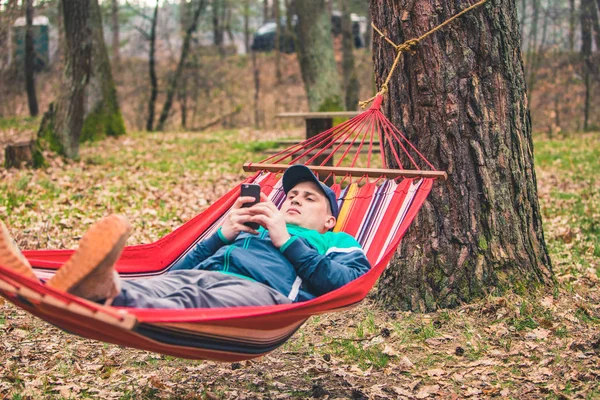 Junger Mann liegt in Hängematte und surft im Handy — Stockfoto