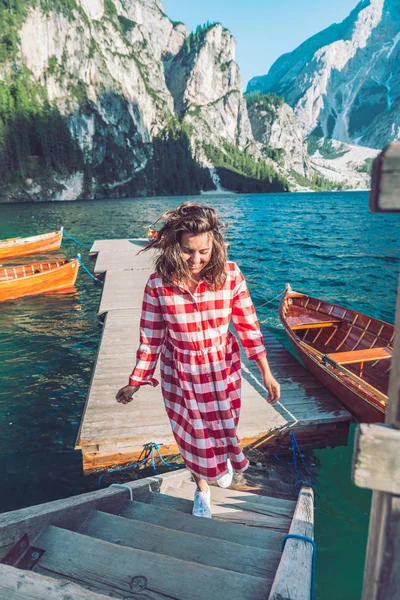 woman walking by mountain lake pier with wooden boats in red dress